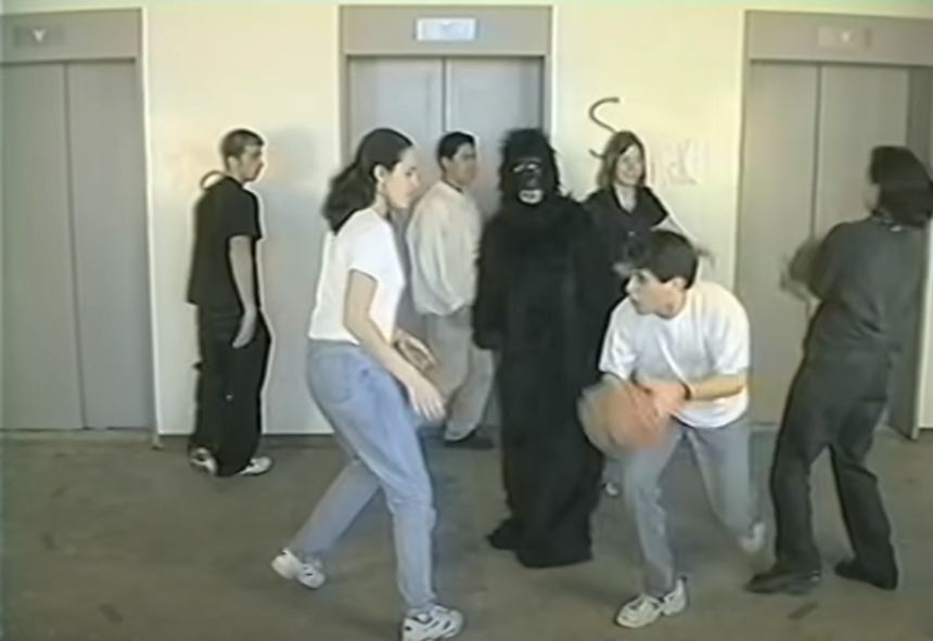 Een scene van een ruimte: op de achtergrond 3 liften. Verspreid door de ruimte staan verscheidene mensen in witte en zwarte t-shirts. Twee mensen in witte t-shirts staan dichtbij de camera, naar elkaar toe gericht. De rechterpersoon is licht door de knieën gebogen en houdt een bruine basketbal vast. In het midden van de ruimte staat een persoon in een gorillapak, kijkend naar de camera.